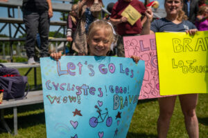 Young girl smiles while holding a light blue poster about bicycle safety at an outdoor rally. Behind her are other sign holders with colorful posters.