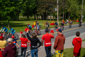 Group of cyclists racing along a paved road beside a grassy park, with spectators cheering on the sidewalk and wearing bright red shirts.