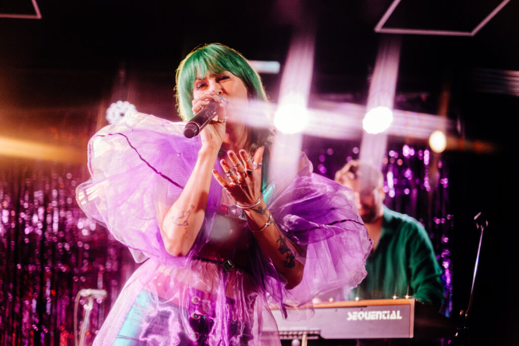 Female singer with green hair performs on stage, pink translucent outfit, microphone in hand, purple sequined backdrop glowing behind.